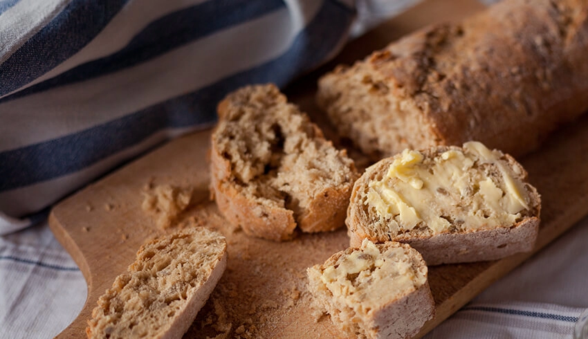 A loaf of bread is cut up on a wooden board. One slice is slathered in butter near another half-eaten. A blue striped kitchen cloth adds to the homemade look.A loaf of bread is cut up on a wooden board. One slice is slathered in butter near another half-eaten. A blue striped kitchen cloth adds to the homemade look.