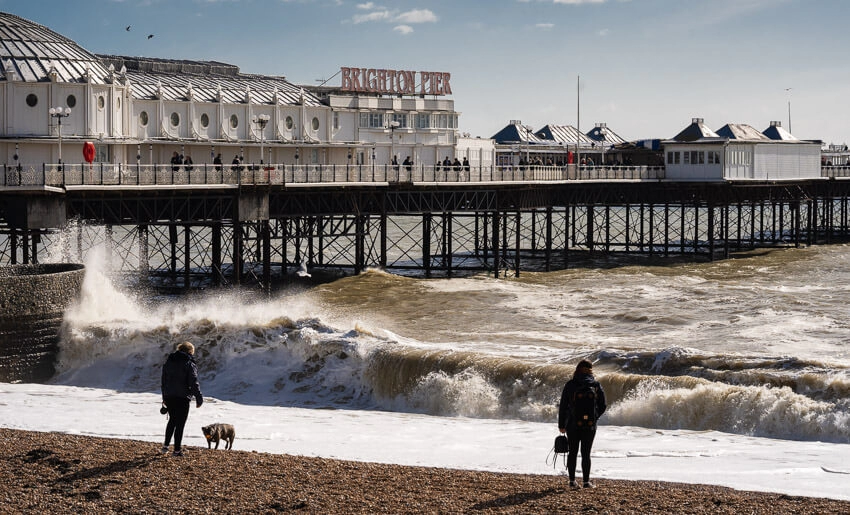 Brighton pier shown with turbulent waves crashing onto the shore. Lots of sea spray and whitewater can be seen as people watch on from the stone beach.