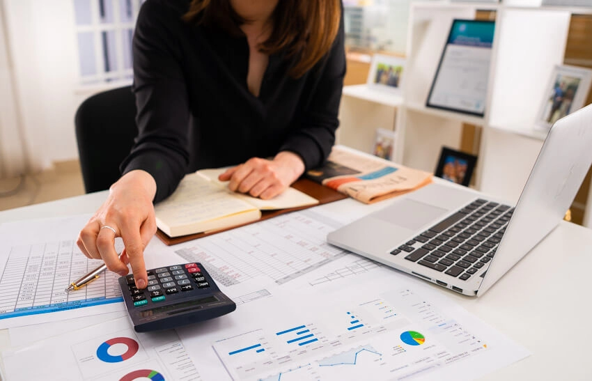 A medium shot of a busy woman sat at a desk working as she types on a calculator. A laptop and multiple papers with charts and a notebook surround her.