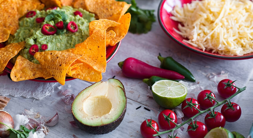 A bowl of nachos with a colourfully contrasting guac dip and chillis are placed near a bowl of cheese. Fresh ingredients including shiny tomatoes are loose.