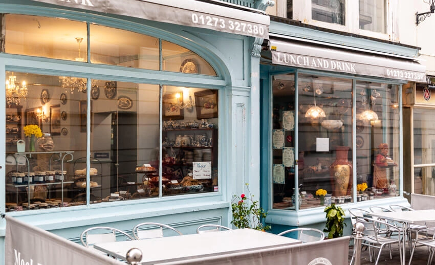 Freshly baked goods and jars can be seen in the front window of an old-fashioned style shop. Bright natural lighting shines on the outside seating area.