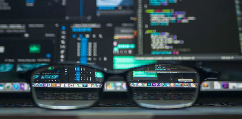 A closeup shot looking through a pair of glasses resting on a computer keyboard. Through the lens a computer screen with code is shown.