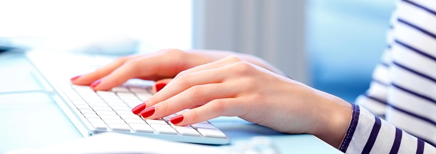 A woman with red painted nails typing onto a Mac desktop keyboard.