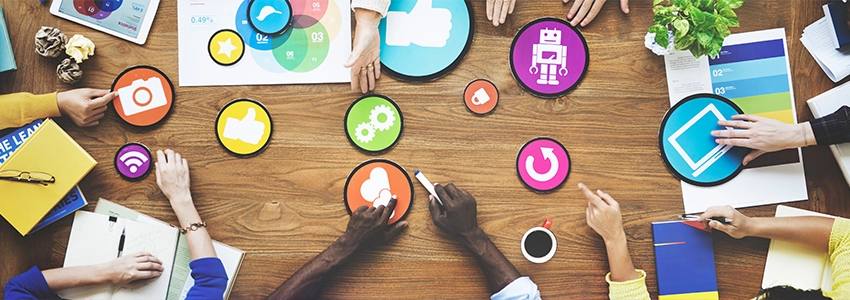 An overhead shot of arms on a table moving cutouts of social media and tech related icons. They are in discussion, some holding stationery near notepads.