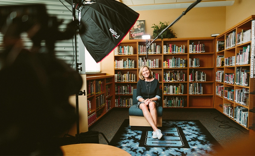 A woman sat in a chair with large bookcases behind her. Video equipment is placed in front of her and she directly faces the camera, while looking up.