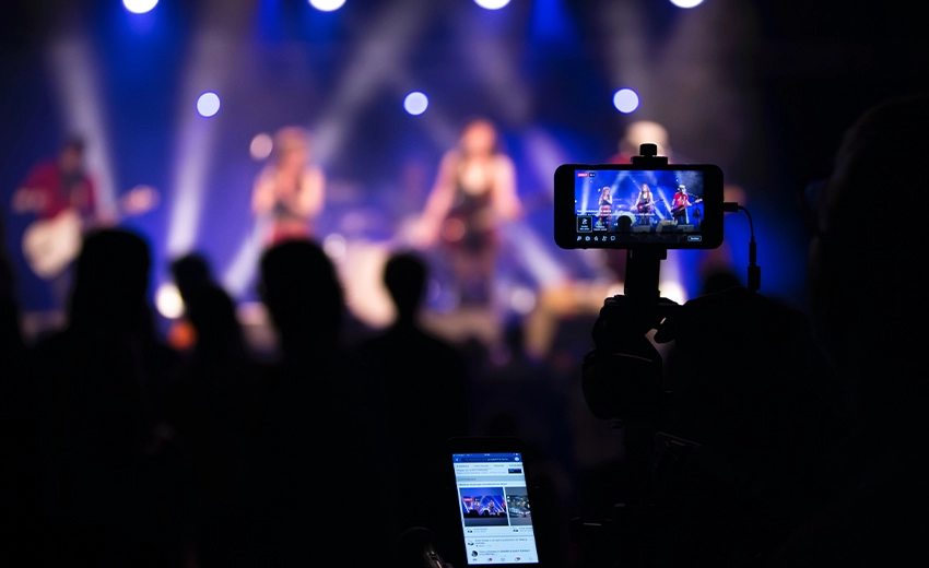 A horizontal mobile screen in focus records a live band blurred in the background of the image. Another phone on social media can be seen in the crowd.