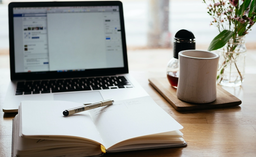 A notebook showing writing is left open with a pen sat on top. It is placed in front of a modern laptop with multiple windows opened.