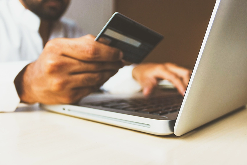 A closeup low angle shot of a man’s hand holding a debit card. The blurred background shows his other hand typing on a laptop keyboard as he looks at the card.