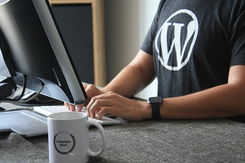 A low angle medium closeup shot of a man’s chest wearing a t-shirt with the WordPress logo. He is sat typing on a keyboard with a monitor in front of him.