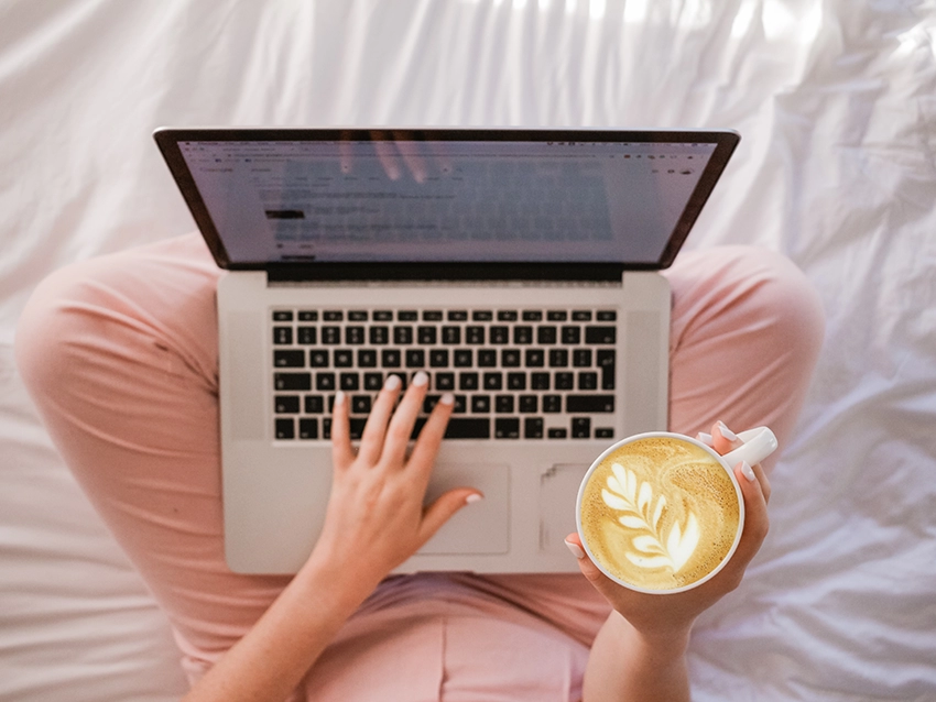 An overhead shot of a laptop balanced on a woman’s lap who is sat on a bed. One hand is placed on the keyboard, while the other holds a mug of coffee.