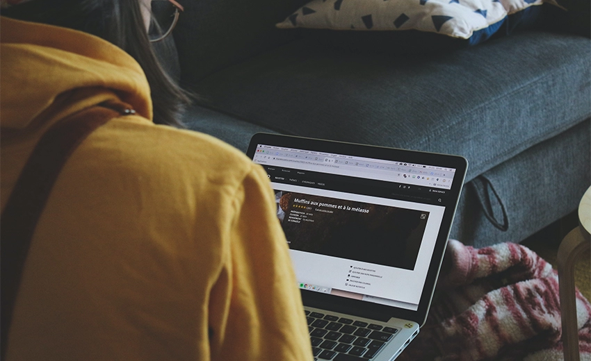 An over the shoulder shot of a woman sat on a dark teal sofa using a laptop.