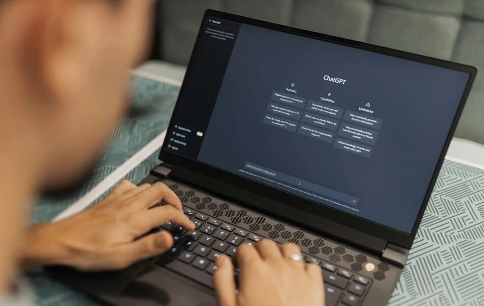 A person sitting at a blue textured table looking at their laptop whilst typing a prompt into OpenAI's ChatGPT generator.