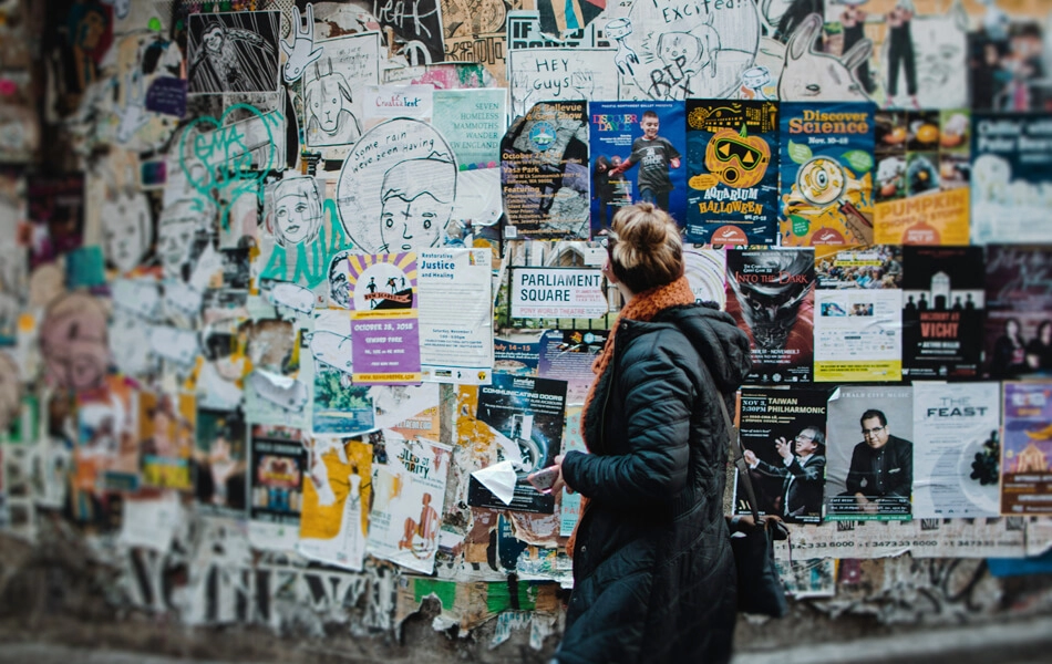 A young woman walking past a brick wall plastered with an array of peeling posters and adverts.