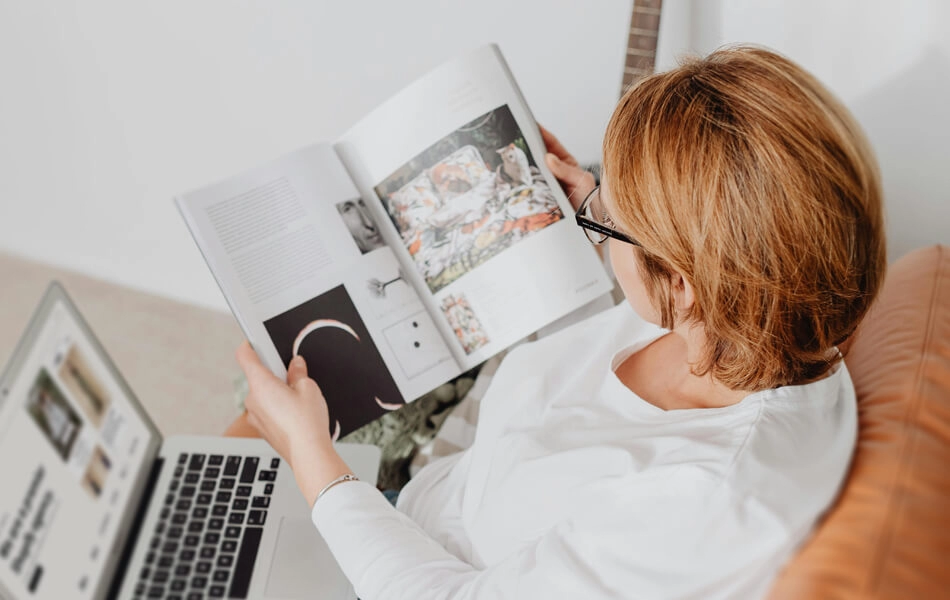 A woman peruses a sleek lifestyle brochure whilst balancing her laptop on her knee.