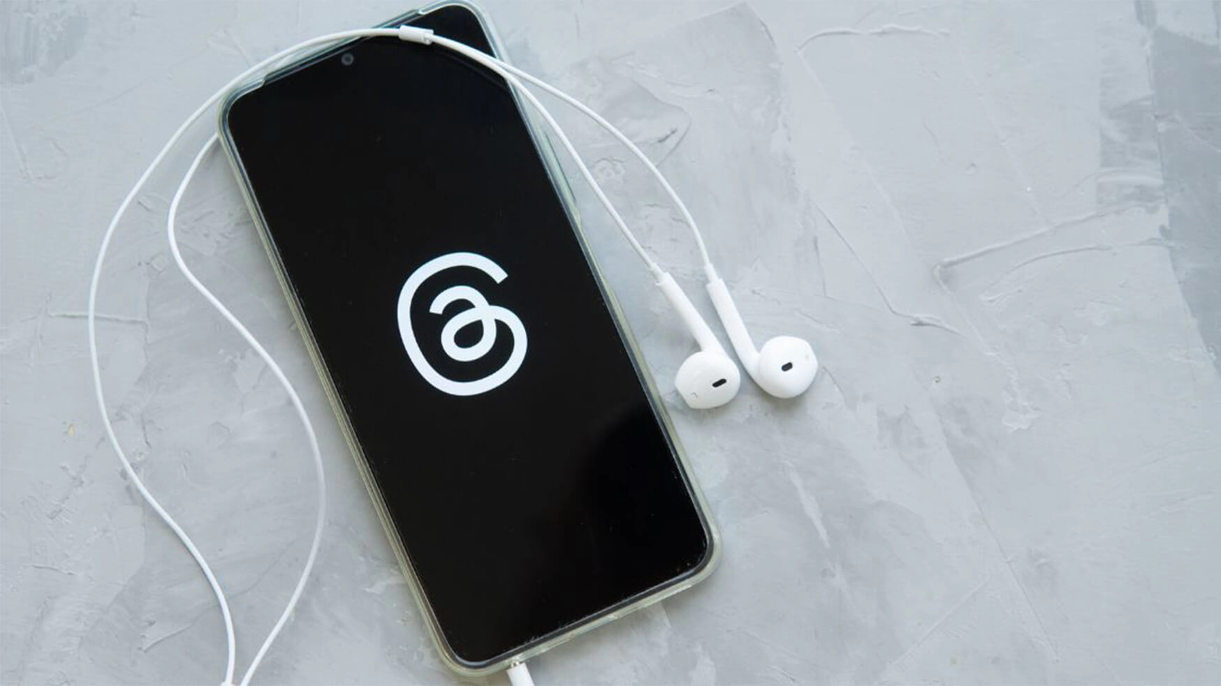 A camera shot looking down at a mobile phone showing the Threads logo. Headphones are plugged in with the wire wrapping round the phone on the desk.