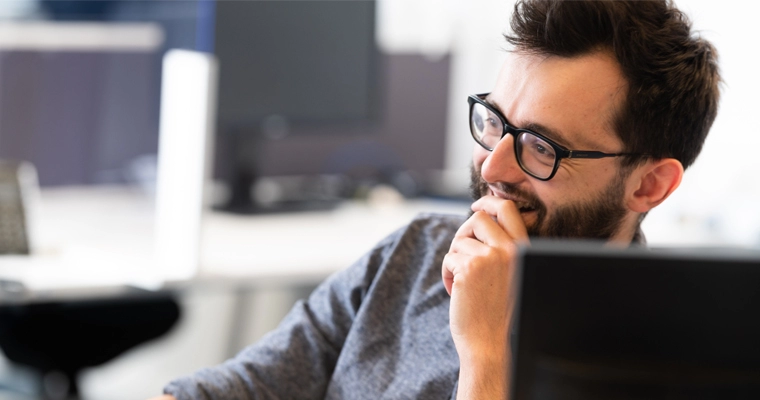 Tim is at his desk and has his hand to his mouth to stifle a laugh. He is wearing a grey sweater and thick black frame glasses.