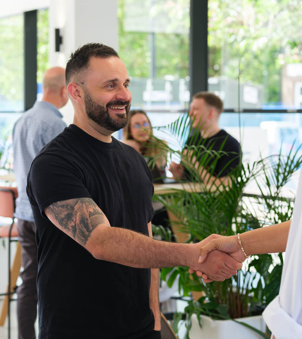 two colleagues shaking hands whilst colleagues work together in the background